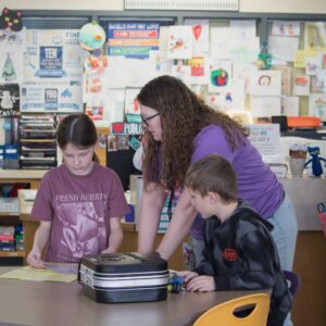 Mrs. Broxtermann gives directions at a table while two students listen.