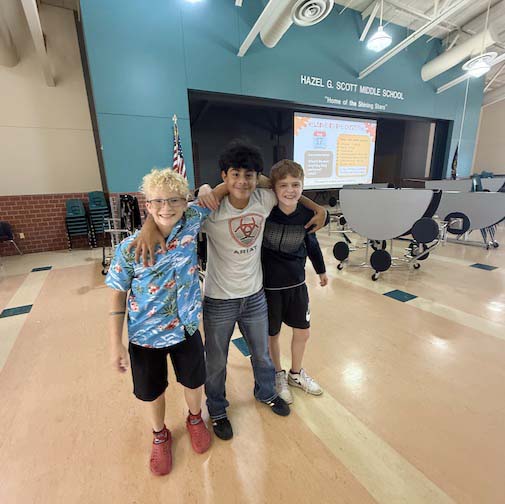 Three Scott Middle School students stand in a row in the middle of the multipurpose room. There are several tables in the background that the students have folded up for custodians.