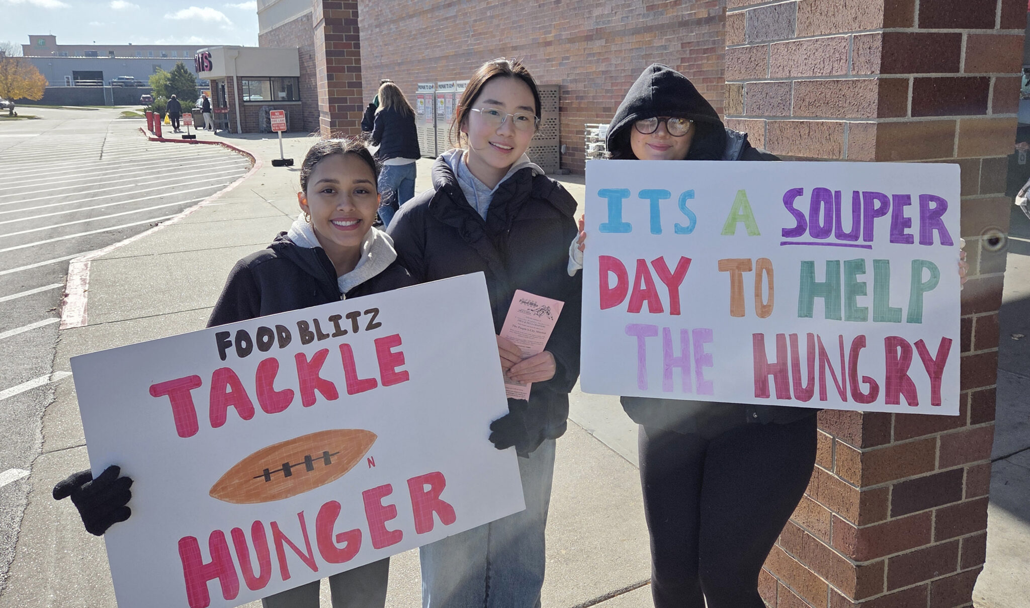 Three Southwest High School students are standing in a row outside a local grocery store for the Food Blitz XIII event. A girl on the left side of the row is holding a sign that says Food Blitz Tackle for Hunger. The girl in the middle is holding a pink delivery card, and the girl on the right is holding a sign that say It's a Souper Day to Help the Hungry.