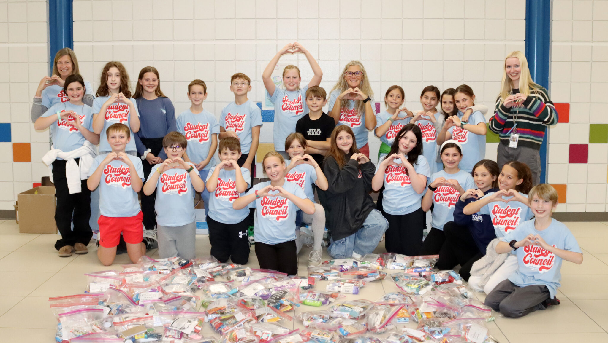 Twenty-two Humann Elementary School Student Council members and three Humann staff members are standing and kneeling in two rows in the front lobby of the school. All of them are forming the shape of a heart with their hands. Many of them are wearing shirts that say Student Council on them. They are behind 123 kindness care kits that they packaged as part of a community service project.