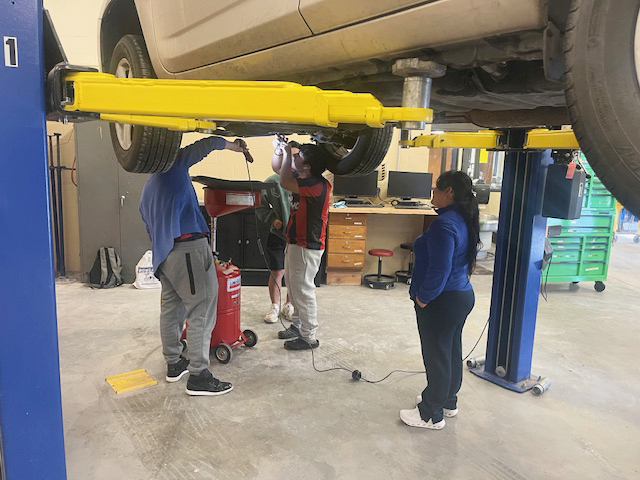 A mother watches as her son and another adult inspect a vehicle in Northeast High School's auto shop. She is standing under the middle of the car, which is raised on a lift. Her son and the other adult are standing in between the front two tires.