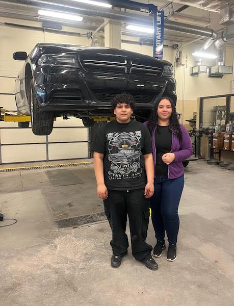 A mother and her son stand in front of a car that is on a lift in the Northeast High School auto shop. The mother has her right arm on the back of her son as they look at the camera.