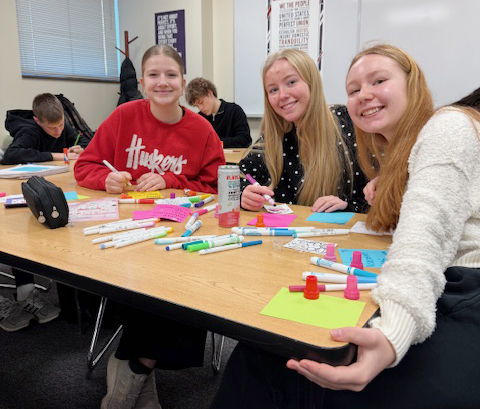 Three Southwest High School students smile at a table in their social studies classroom. The three girls are using markers and pens to write Valentine's Day messages for Lakeview Elementary School students and teachers.