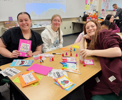 Three Southwest High School students smile at a table in their social studies classroom. They are using markers and glue stick to make Valentine's Day cards for Lakeview Elementary School students and teachers. One of the students is holding a card that says "Teaching can change the world."