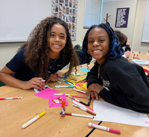 Two Southwest High School students smile at one of their tables in a social studies classroom. They are using pens and markers to make Valentine's Day cards for Lakeview Elementary School students and staff.