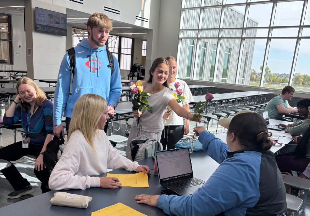 A Standing Bear Hope Squad member hands a rose to a staff member who is sitting at a table in the lunchroom. The student has several other roses in her right hand that she will give to other teachers. A fellow Hope Squad member is standing next to her, and two Standing Bear students and one staff member are on the lefthand side of the photo.