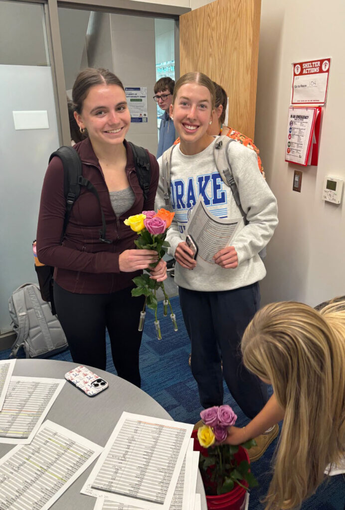 Two Standing Bear Hope Squad members stand in an office. One student is holding a bouquet of roses and the other student is holding a list of classrooms they will visit. They passed out roses to staff members as part of Teacher Appreciation Week.