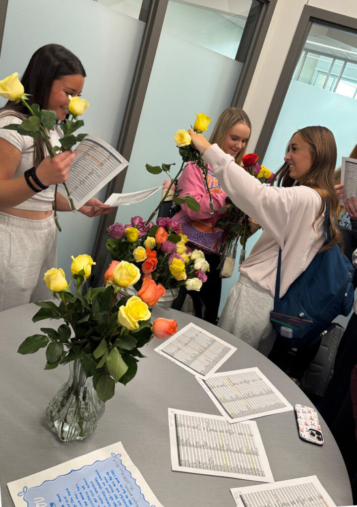 Three Standing Bear Hope Squad members arrange roses in a meeting space at school. One student is holding roses and a list of classrooms they will visit, and two other students are holding several roses in their hands. The three girls took part in a Hope Squad activity for Teacher Appreciation Week.