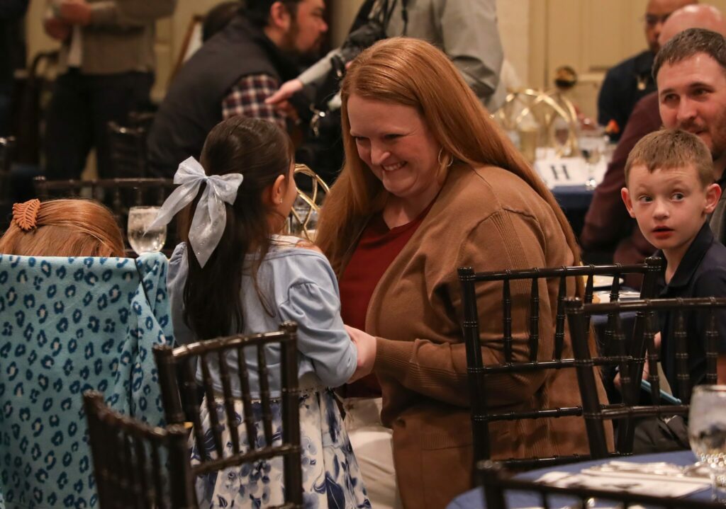 A smiling woman with long auburn hair warmly greets a young girl with a light blue bow in her hair, holding her hands.