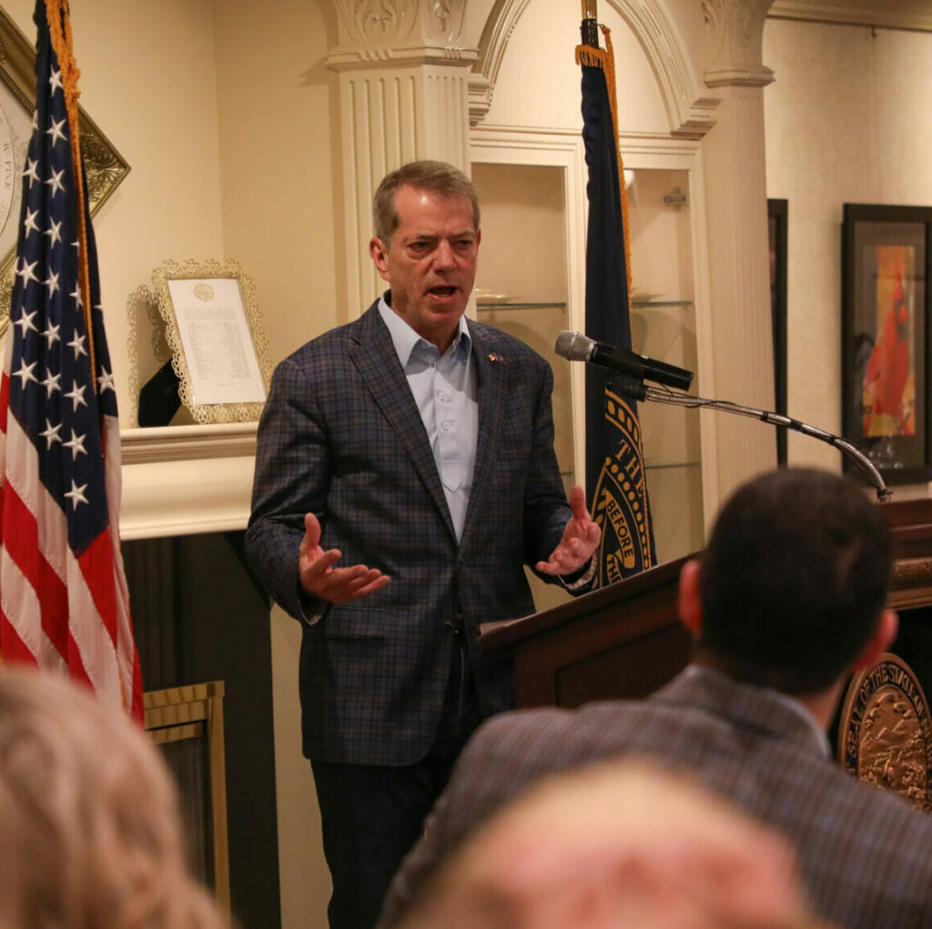 The governor of Nebraska in a plaid suit and blue shirt speaks animatedly at a podium, gesturing with both hands. An American flag is to his left, and the Nebraska state flag is to his right. He addresses the ceremony attendees.