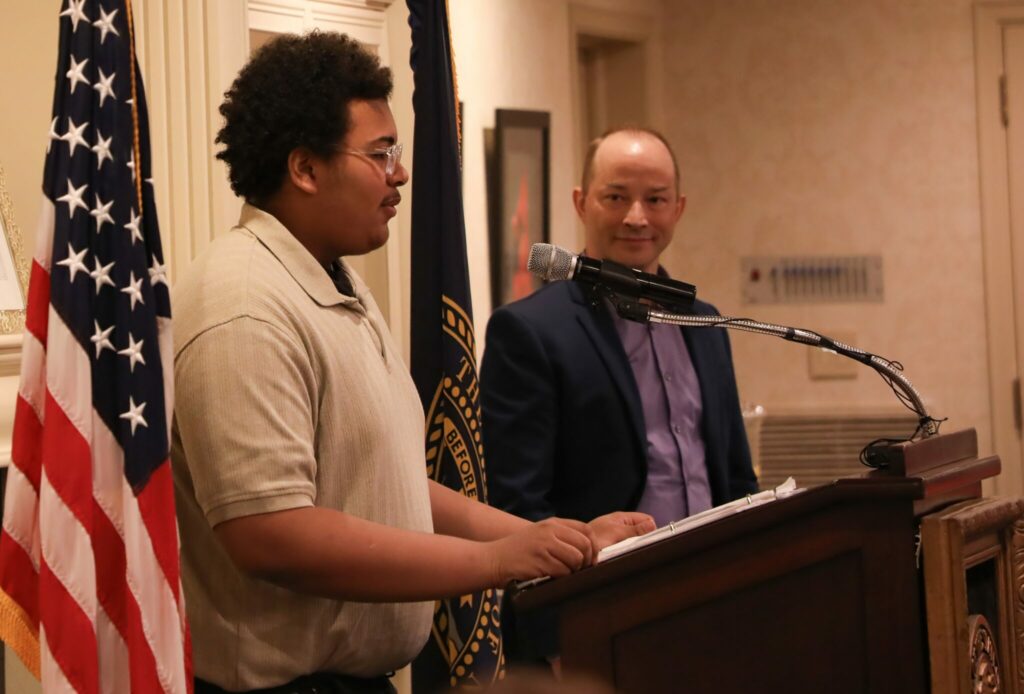 A middle school student stands at a podium, speaking into a microphone while a teacher stands to his side, watching