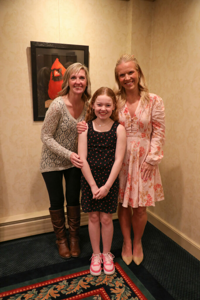 Three smiling females, two adults and one young girl, stand together for a photo indoors. The adults wear a patterned sweater and a floral dress, while the girl wears a black polka-dot dress and pink sneakers. A framed picture of a red cardinal hangs behind them.