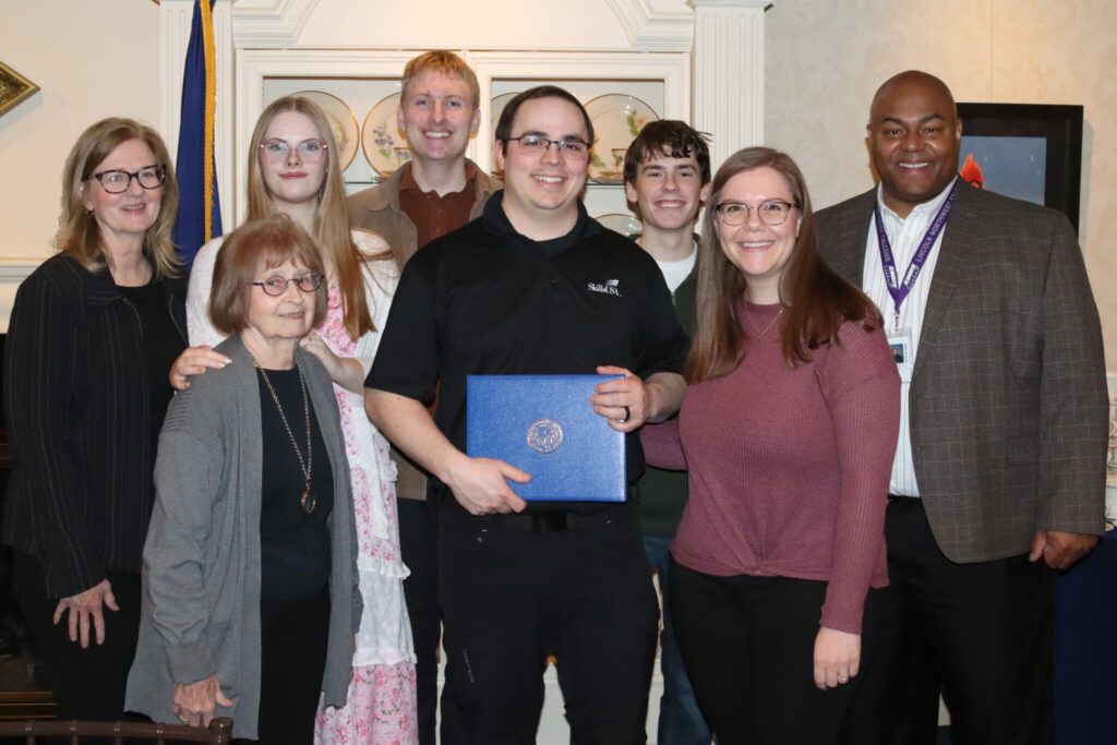 Nine smiling people, six women and three men, pose indoors. A man in the center wears a black shirt and proudly holds a blue award folder.