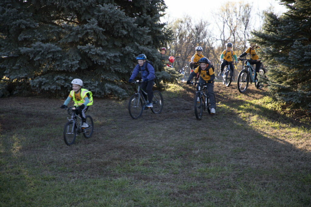 Students having fun riding bicycles down a gentle grass-covered hill between two pine trees