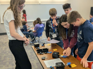 A high school student and four middle school students stand around a table working on an activity.