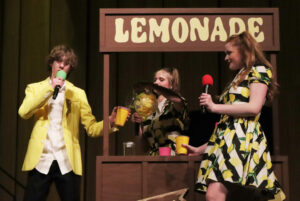 Show choir event with girl in lemonade stand pouring lemonade into a boy's cup while another girl watches