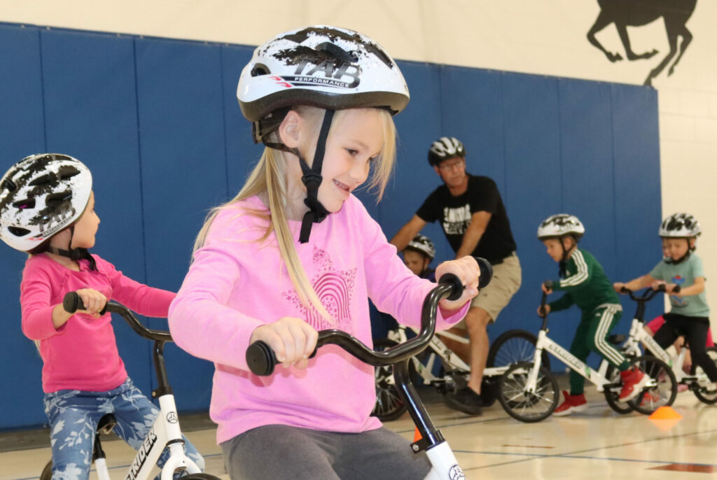 An elementary aged girl in a pink shirt and bike helmet rides on a bike in a gymnasium