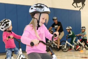 An elementary aged girl in a pink shirt and bike helmet rides on a bike in a gymnasium