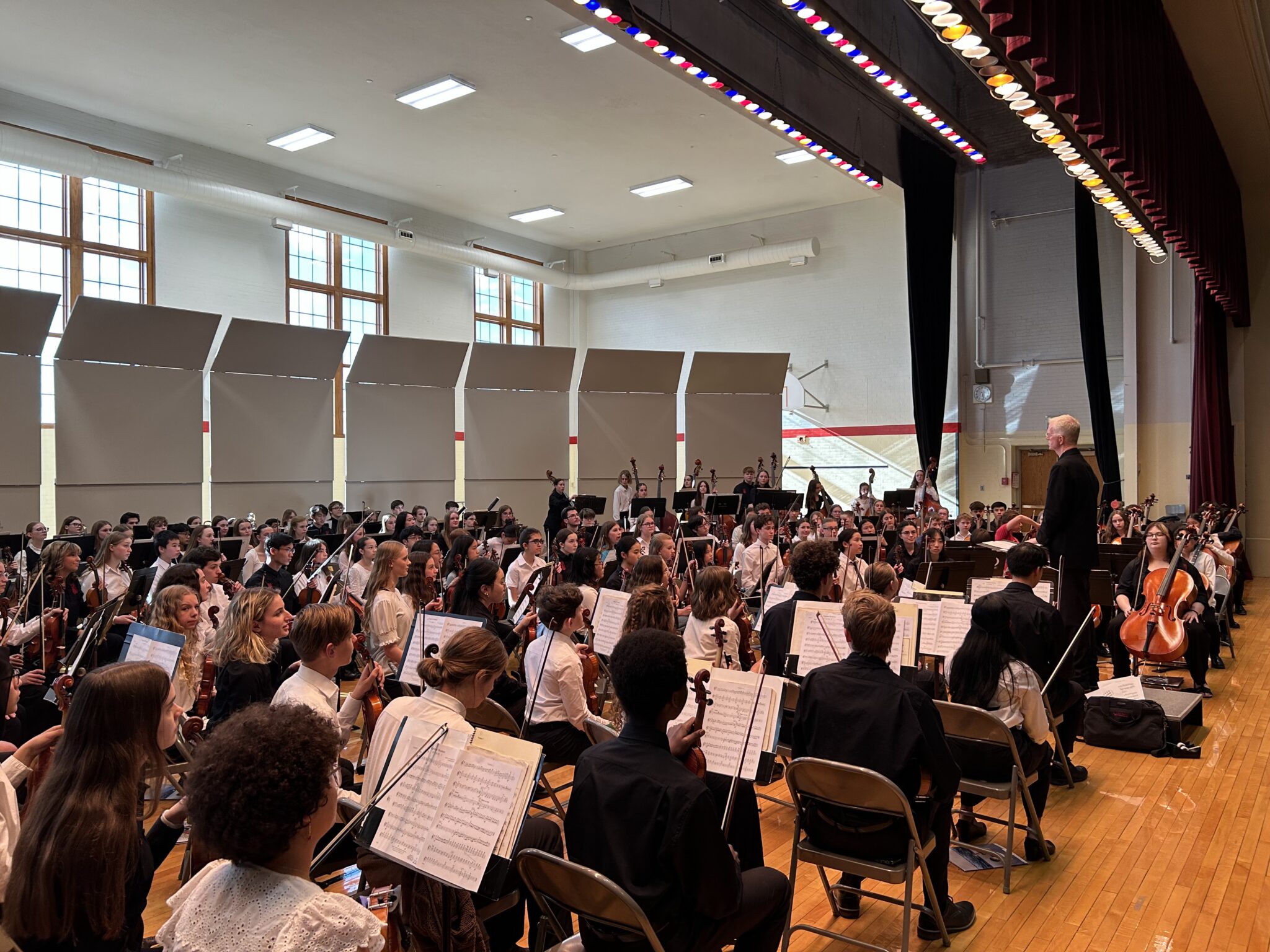 A large group of students site, prepared to play as their conductor stands at the center.